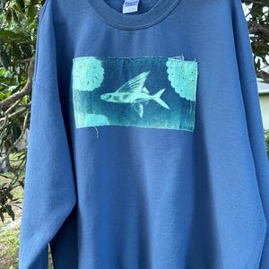 May include: A blue crewneck sweatshirt featuring a rectangular patch on the chest. The patch displays a light green flying fish graphic centered between two circular lace-like patterns, with raw, frayed edges and the word INDIGO printed in the corner.