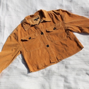 May include: A mustard yellow corduroy jacket featuring a collared neckline, a front button-down closure, and two chest pockets with buttoned flaps. The long sleeves have buttoned cuffs, and the garment has a cropped length and textured fabric.