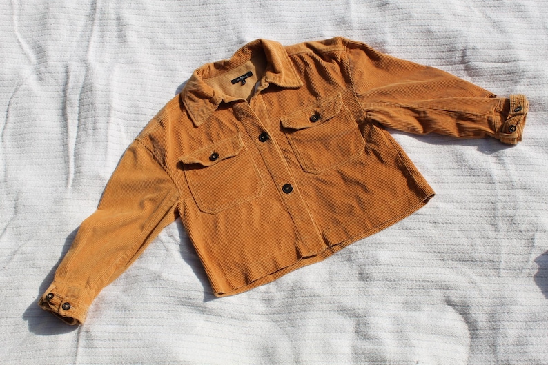 May include: A mustard yellow corduroy jacket featuring a collared neckline, a front button-down closure, and two chest pockets with buttoned flaps. The long sleeves have buttoned cuffs, and the garment has a cropped length and textured fabric.