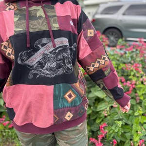 May include: A patchwork hoodie in shades of burgundy, black, and olive green. The front features a graphic of a winged creature with text. The sleeves and body are made of various patterned fabrics. The hoodie has a drawstring and a kangaroo pocket.