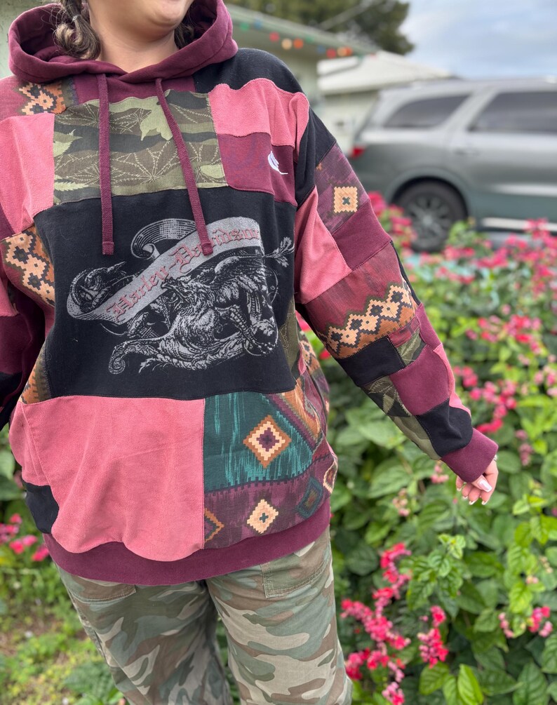 May include: A patchwork hoodie in shades of burgundy, black, and olive green. The front features a graphic of a winged creature with text. The sleeves and body are made of various patterned fabrics. The hoodie has a drawstring and a kangaroo pocket.