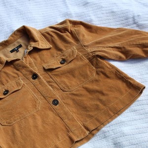 May include: A mustard yellow corduroy jacket featuring a collared neckline, a front button closure, and two chest pockets with buttoned flaps. The long sleeves have buttoned cuffs, and the interior label displays the 7 For All Mankind brand logo.