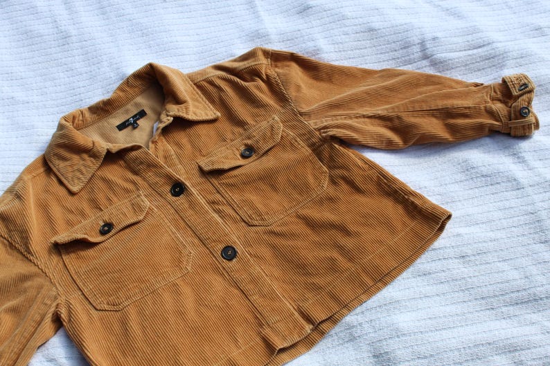 May include: A mustard yellow corduroy jacket featuring a collared neckline, a front button closure, and two chest pockets with buttoned flaps. The long sleeves have buttoned cuffs, and the interior label displays the 7 For All Mankind brand logo.