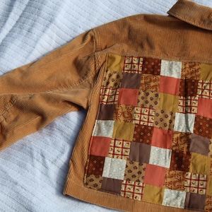May include: A tan corduroy jacket featuring a large back panel constructed from a patchwork of various fabric squares in shades of brown, orange, cream, and mustard yellow, with a standard collar and long sleeves with buttoned cuffs.