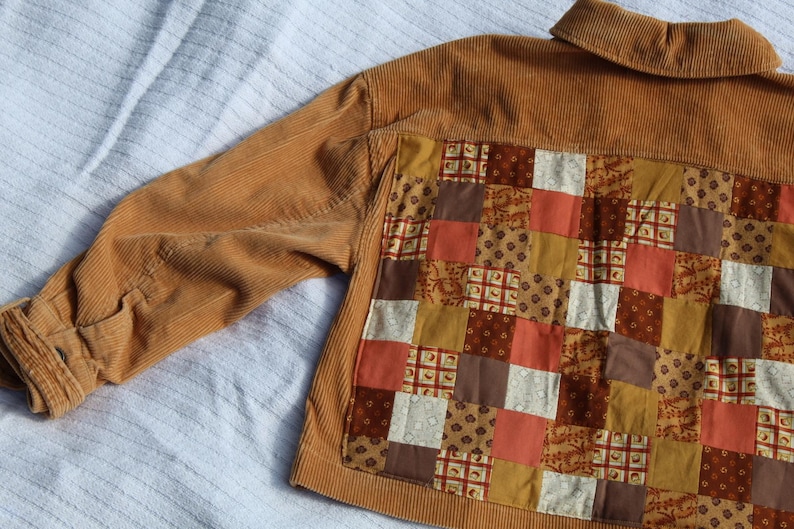 May include: A tan corduroy jacket featuring a large back panel constructed from a patchwork of various fabric squares in shades of brown, orange, cream, and mustard yellow, with a standard collar and long sleeves with buttoned cuffs.