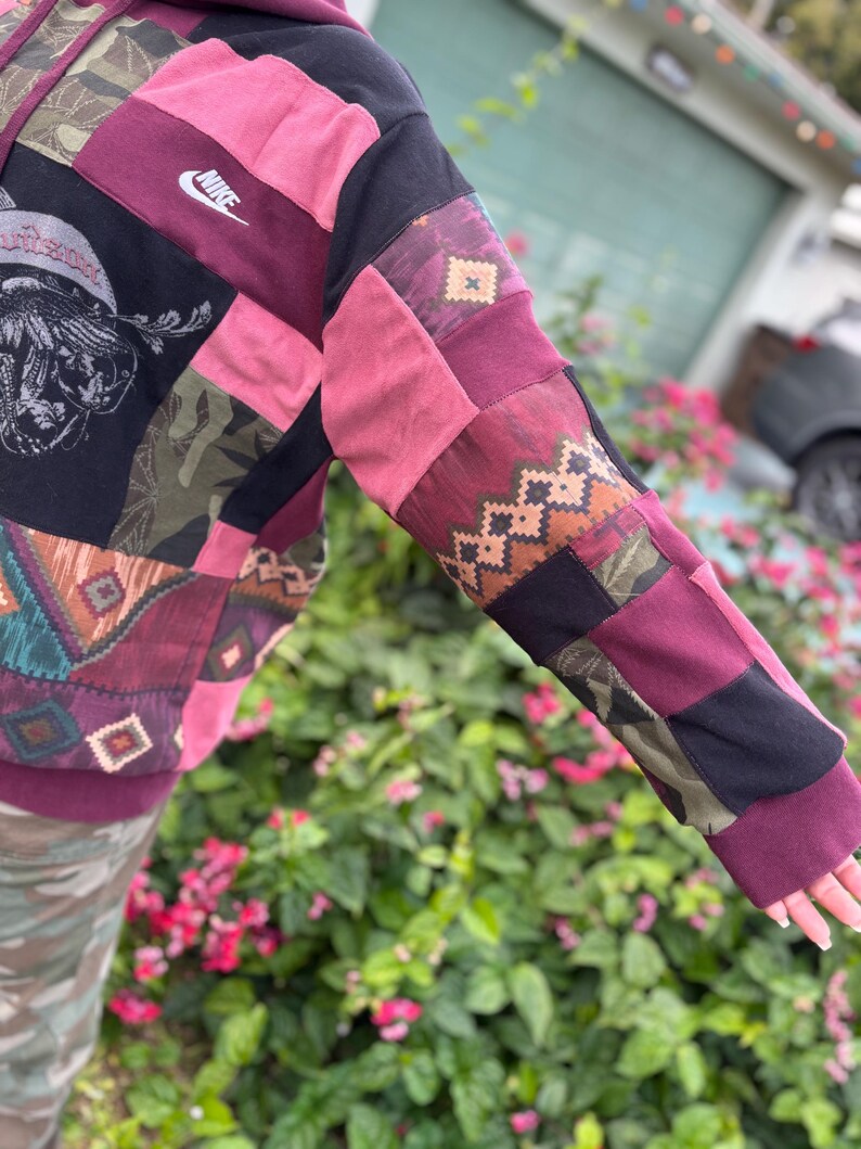 May include: A patchwork hoodie with a Nike logo. The sleeves and body are made of various fabrics in shades of burgundy, black, pink, and olive green. The design includes geometric and tribal patterns.
