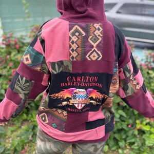 May include: A patchwork hoodie in shades of pink, burgundy, and black. The back features a large graphic with the text "CARLTON HARLEY-DAVIDSON" and "AUTHENTIC MANTUA, OHIO". The design incorporates various patterns and textures.