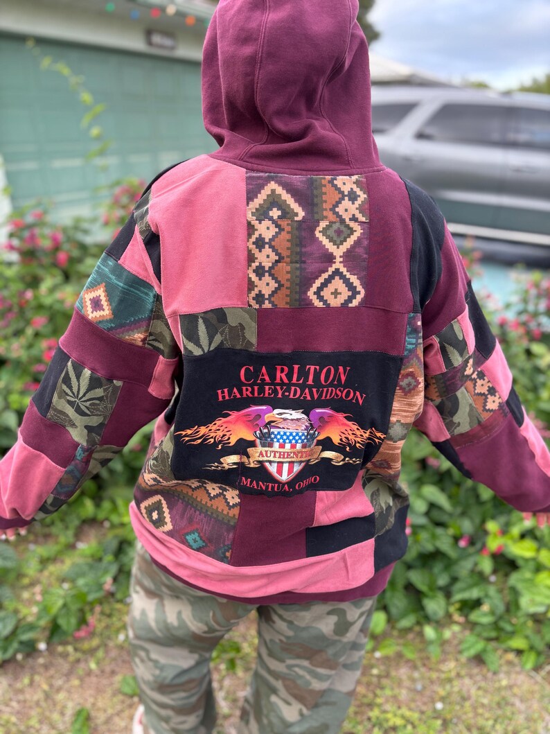 May include: A patchwork hoodie in shades of pink, burgundy, and black. The back features a large graphic with the text "CARLTON HARLEY-DAVIDSON" and "AUTHENTIC MANTUA, OHIO". The design incorporates various patterns and textures.