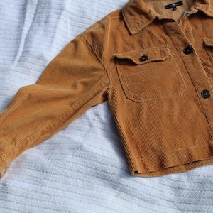 May include: A mustard yellow corduroy jacket featuring a collared neckline, long sleeves with buttoned cuffs, and two chest pockets with button-down flaps. The front closure consists of dark buttons, and the interior label reads 7 For All Mankind.