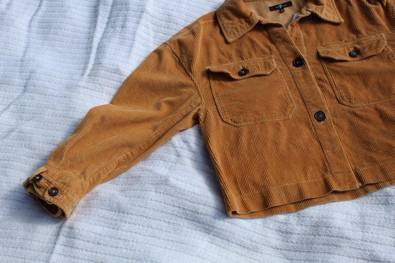 May include: A mustard yellow corduroy jacket featuring a collared neckline, long sleeves with buttoned cuffs, and two chest pockets with button-down flaps. The front closure consists of dark buttons, and the interior label reads 7 For All Mankind.