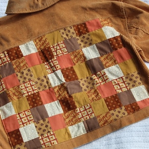May include: A tan corduroy jacket featuring a large rectangular back panel composed of a patchwork design. The patchwork consists of various square fabric pieces in shades of brown, orange, mustard yellow, and cream, featuring floral and geometric patterns.
