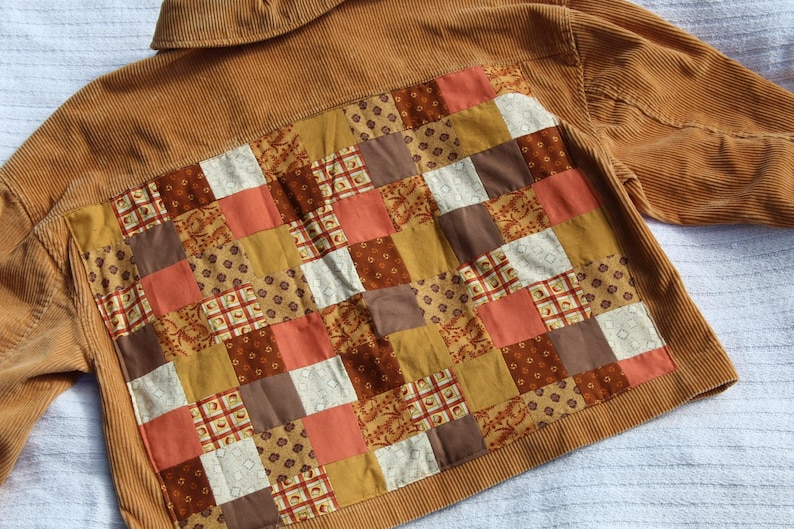 May include: A tan corduroy jacket featuring a large rectangular back panel composed of a patchwork design. The patchwork consists of various square fabric pieces in shades of brown, orange, mustard yellow, and cream, featuring floral and geometric patterns.