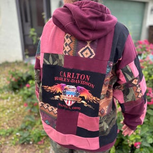 May include: A patchwork hoodie in shades of burgundy, pink, and black. The back features a Harley-Davidson logo with the text "CARLTON HARLEY-DAVIDSON AUTHENTIC MANTUA, OHIO". The hoodie has a hood and is made of various fabric patterns.