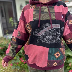 May include: A patchwork hoodie in burgundy, black, and pink with a Harley-Davidson graphic. The hoodie features a drawstring hood and a kangaroo pocket. The design incorporates various fabric patterns, including camouflage and geometric shapes.