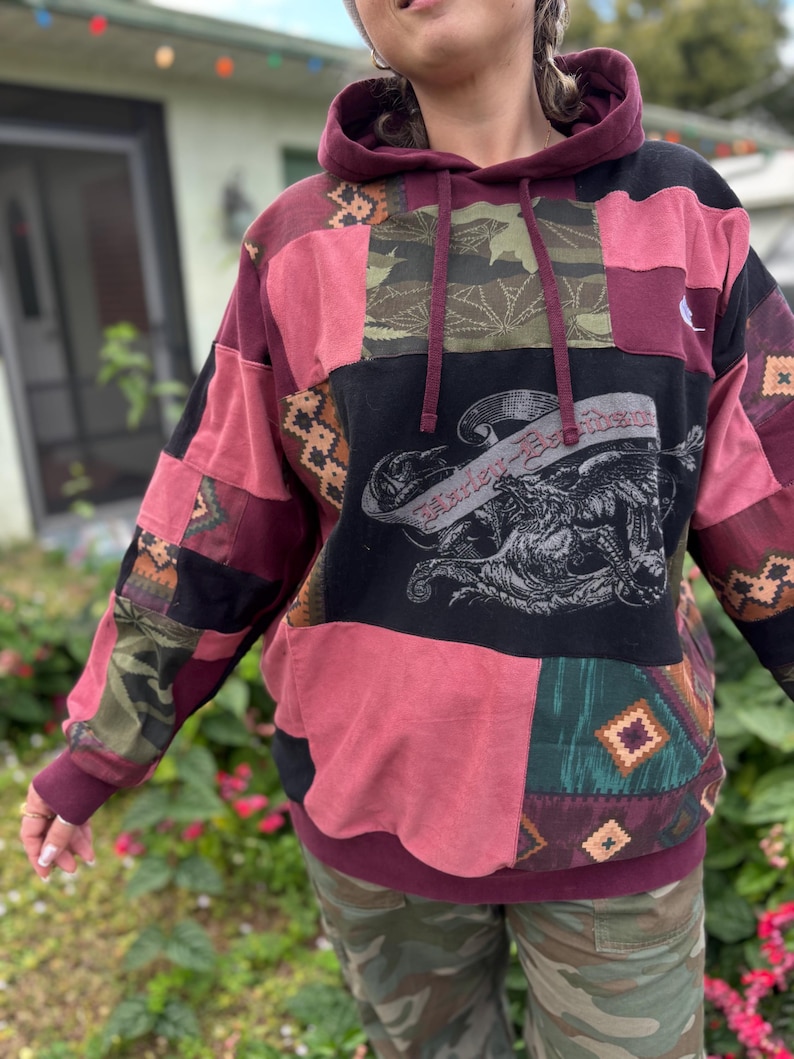 May include: A patchwork hoodie in burgundy, black, and pink with a Harley-Davidson graphic. The hoodie features a drawstring hood and a kangaroo pocket. The design incorporates various fabric patterns, including camouflage and geometric shapes.