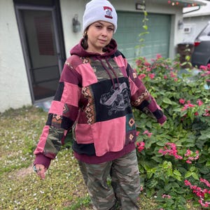 May include: A patchwork hoodie in shades of pink, burgundy, and black, with a graphic design on the front. The hoodie is paired with camouflage pants and a white beanie with a graphic.