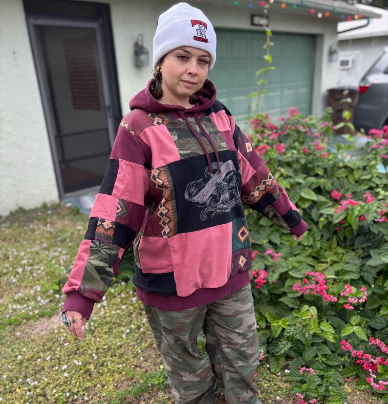 May include: A patchwork hoodie in shades of pink, burgundy, and black, with a graphic design on the front. The hoodie is paired with camouflage pants and a white beanie with a graphic.
