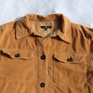 May include: A mustard yellow corduroy shirt featuring a classic collar, a front button-down closure with dark buttons, and two chest pockets with buttoned flaps. The interior neck label displays the text 7 For All Mankind with a size M.