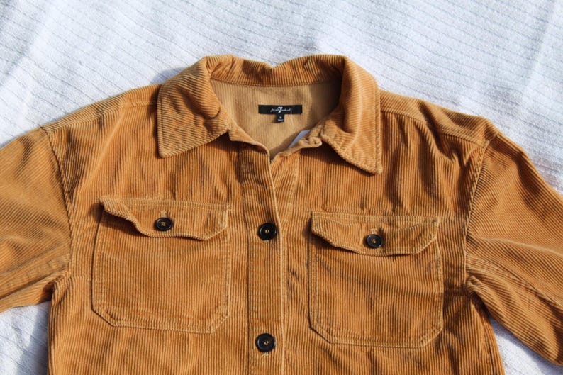 May include: A mustard yellow corduroy shirt featuring a classic collar, a front button-down closure with dark buttons, and two chest pockets with buttoned flaps. The interior neck label displays the text 7 For All Mankind with a size M.