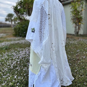 May include: A white, layered garment with lace and cut-out details, draped on a mannequin. The garment appears to be made from repurposed textiles, with visible stitching and varying textures. A light green and yellow fabric is visible.