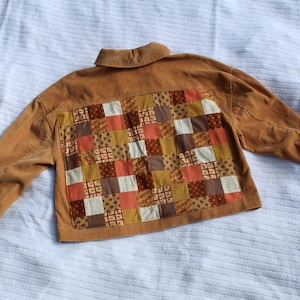 May include: A tan corduroy jacket featuring a large rectangular back panel composed of a patchwork grid of various fabric squares in shades of brown, orange, cream, and gold, with a standard collar and long sleeves with buttoned cuffs.