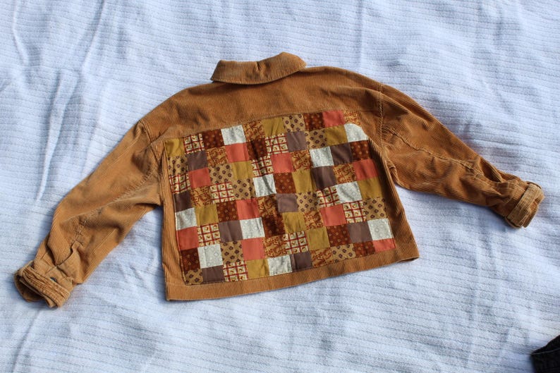 May include: A tan corduroy jacket featuring a large rectangular back panel composed of a patchwork grid of various fabric squares in shades of brown, orange, cream, and gold, with a standard collar and long sleeves with buttoned cuffs.