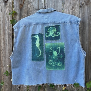 May include: A light blue denim vest featuring three rectangular fabric patches on the back. The patches display green illustrations of a seahorse, a jellyfish, and an octopus. The vest has a collared neckline and a frayed, raw-cut bottom hem.