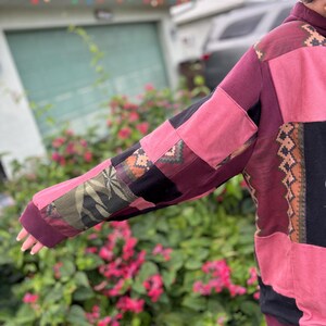 May include: A patchwork hoodie in shades of pink, burgundy, and black. The sleeves feature a variety of patterned fabric patches, including a leaf design. The hoodie has a drawstring hood and a relaxed fit, perfect for casual wear.