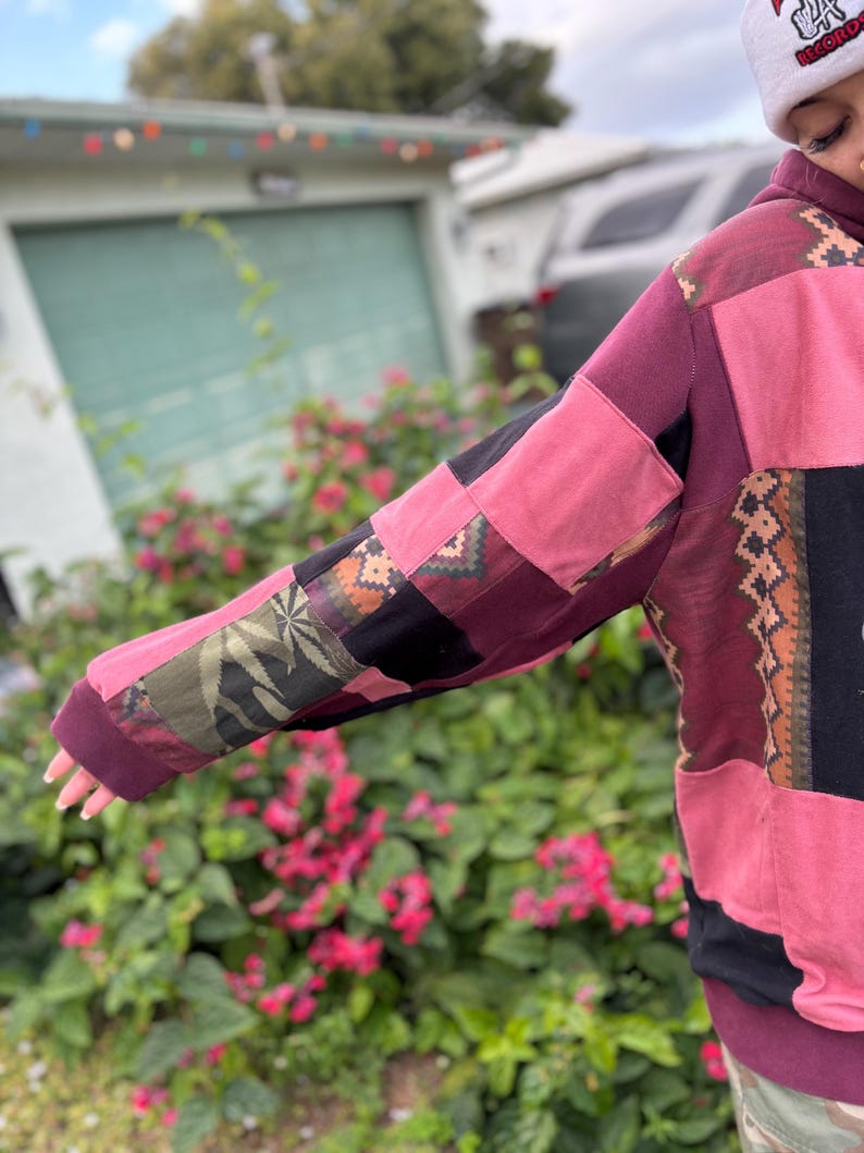 May include: A patchwork hoodie in shades of pink, burgundy, and black. The sleeves feature a variety of patterned fabric patches, including a leaf design. The hoodie has a drawstring hood and a relaxed fit, perfect for casual wear.