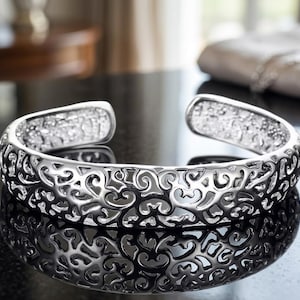 May include: A silver cuff bracelet with an open design. The bracelet features an intricate, swirling pattern throughout its surface. The metal reflects light, creating a bright and detailed appearance. The bracelet is displayed on a dark, reflective surface.