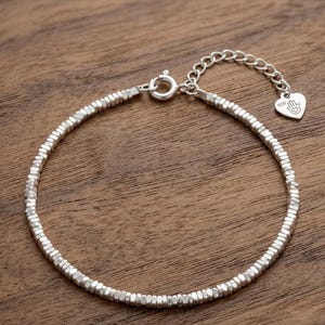 May include: A silver bracelet with a beaded design. The bracelet features small, faceted beads strung together, with a heart-shaped charm engraved with a hand symbol. The bracelet is on a wooden surface.