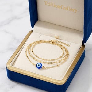 May include: A gold-coloured layered bracelet set with a blue evil eye charm, presented in an open blue velvet box. The bracelets are displayed against a cream-coloured interior. The brand name "TrillionGallery" is embossed in gold.