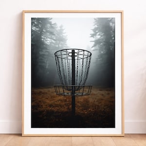 May include: A framed photograph of a disc golf basket in a foggy forest. The black metal basket is centered, with chains hanging down. The background features tall trees and a misty atmosphere, creating a serene outdoor scene.