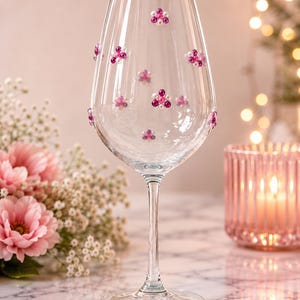 Op de afbeelding: Een wijnglas van helder glas versierd met kleine clusters van roze en witte kralen, gerangschikt in bloemmotieven over de kelk. Het glas heeft een slanke steel en een ronde voet, waarbij de kraalclusters over het oppervlak zijn verspreid.