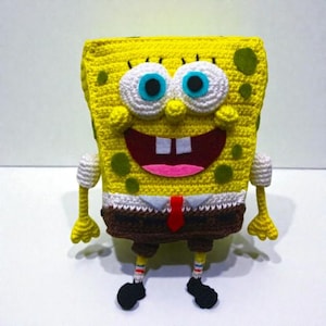 Könnte beinhalten: Eine handgefertigte Häkelfigur einer bekannten Zeichentrickfigur. Die Figur ist gelb mit braunen Shorts, einem weißen Hemd und einer roten Krawatte. Sie hat große weiße Augen mit blauen Pupillen, ein breites Lächeln und schwarze Schuhe. Die Figur steht aufrecht.