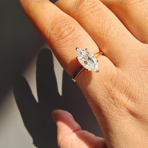 May include: A marquise-cut diamond engagement ring with a gold band. The diamond is set with four prongs and sparkles in the sunlight. The ring is shown on a person's finger.