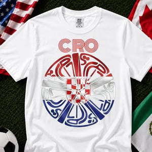 May include: A white short-sleeve t-shirt featuring a circular graphic design with the text CRO at the top. The center of the circle displays the red and white checkered pattern of the Croatian flag, surrounded by stylized blue and red line art.