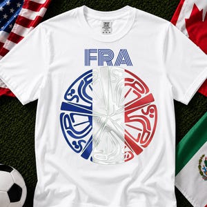 May include: A white short-sleeve t-shirt featuring the text FRA in blue stripes above a circular graphic. The circle is divided into vertical blue, white, and red sections with stylized, abstract line art patterns integrated throughout the design.