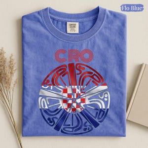 May include: A Flo Blue short-sleeve t-shirt featuring a circular graphic design with the text CRO above it. The graphic incorporates a red and white checkered pattern within a stylized circular shape, layered over red, white, and blue horizontal stripes.