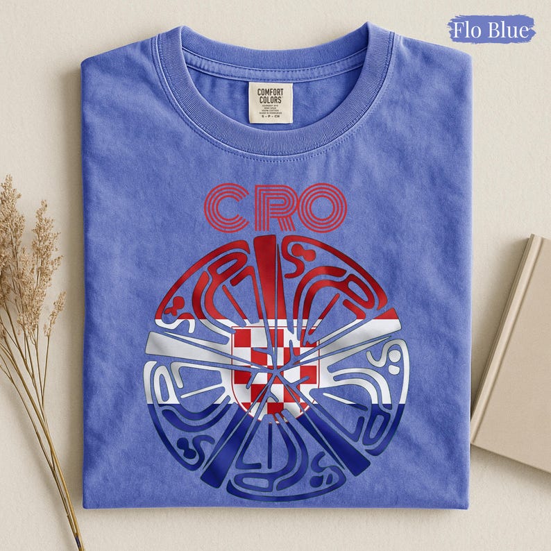 May include: A Flo Blue short-sleeve t-shirt featuring a circular graphic design with the text CRO above it. The graphic incorporates a red and white checkered pattern within a stylized circular shape, layered over red, white, and blue horizontal stripes.