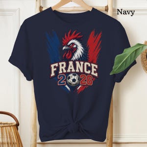 May include: Navy blue t-shirt featuring a rooster design with red, white, and blue accents, and the text "FRANCE 2026" with a soccer ball graphic. The shirt is a short-sleeved crew neck.