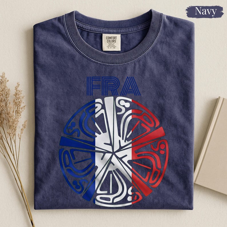 May include: A navy blue crew neck t-shirt featuring the text FRA in blue outline letters above a circular graphic. The circular design incorporates the blue, white, and red colors of the French flag with intricate, stylized, and abstract line patterns.