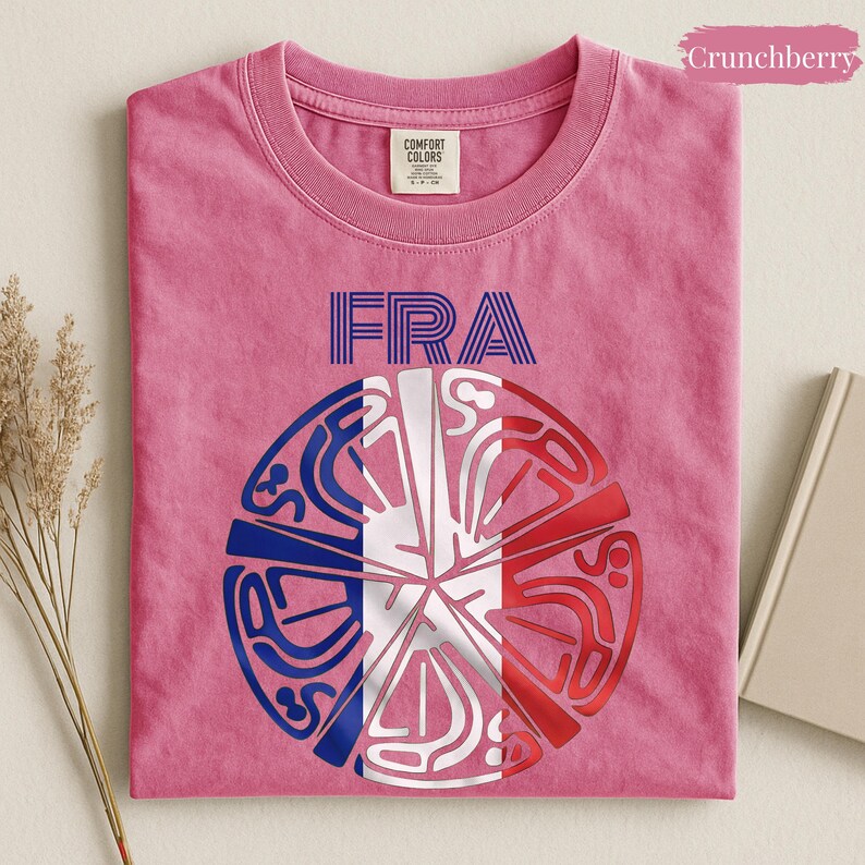 May include: A pink short-sleeve t-shirt featuring the text FRA in blue and a circular graphic design below it. The graphic consists of blue, white, and red segments arranged in a radial pattern, resembling the French flag colors with intricate lines.