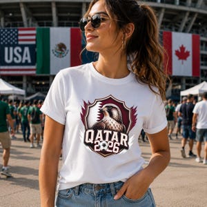 May include: White t-shirt featuring a maroon and white crest with a falcon and the text "QATAR 2026". The shirt is worn by a person in front of flags of the USA, Mexico, and Canada.