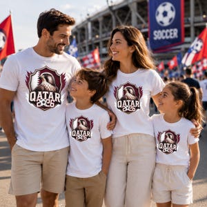 May include: A family of four wearing matching white t-shirts with a Qatar 2026 design. The shirts feature a maroon and white crest with a falcon and the text "QATAR 2026". The family is standing in front of a stadium with soccer flags.