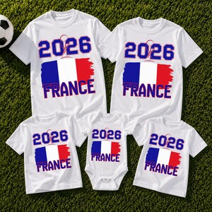 May include: White t-shirts and a onesie with a French flag design. The shirts and onesie have the text "2026 FRANCE" in blue and red. A soccer ball is visible in the background.