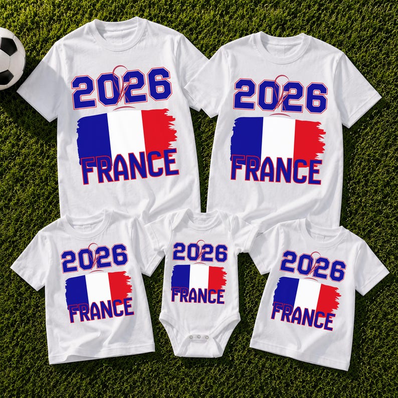 May include: White t-shirts and a onesie with a French flag design. The shirts and onesie have the text "2026 FRANCE" in blue and red. A soccer ball is visible in the background.