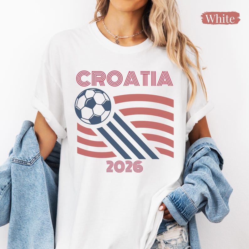 May include: A white t-shirt featuring a graphic design with the text CROATIA and 2026 in red, accompanied by a soccer ball illustration and red and blue wavy lines. The shirt is worn with a blue denim jacket draped over the shoulders.
