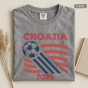May include: A gray short-sleeve t-shirt featuring the text CROATIA in red, a graphic of a soccer ball, and the year 2026 in red. The design includes red and dark blue stripes. The shirt has a Comfort Colors brand label at the inner neckline.