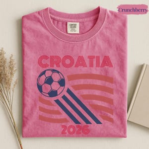 May include: A pink short-sleeved t-shirt featuring the text CROATIA in red, a dark blue soccer ball graphic with motion lines, and the year 2026 in red. The shirt has a Comfort Colors brand label on the inside of the collar and a pink tag.